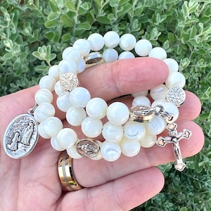 May include: A stretch bracelet composed of round, iridescent white beads featuring several silver-toned metal spacers with religious imagery, a dangling oval medal depicting a kneeling figure, and a silver-toned crucifix pendant.