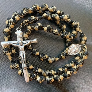 May include: A black and tan camouflage paracord rosary with a silver crucifix and a silver medal featuring a depiction of the Virgin Mary and child. The crucifix has the inscription "INRI" on the crossbar.
