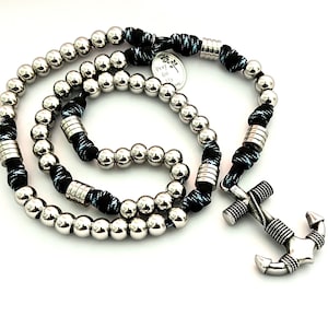 Könnte beinhalten: Ein silberner Rosenkranz mit nautischem Thema. Der Rosenkranz hat glänzende, runde Perlen, eine schwarz-weiße Kordel und einen silbernen Ankeranhänger. Ein kleiner, runder Anhänger mit dem Text "Pray for us" ist ebenfalls vorhanden.