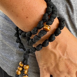 Op de afbeelding: Zwart paracord armband met bruine tijgeroog kralen. De armband is geknoopt en heeft een kwast met extra tijgeroog kralen.