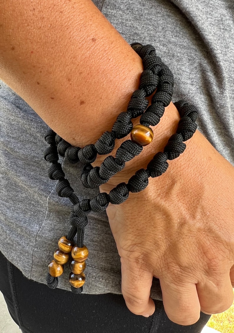 Op de afbeelding: Zwart paracord armband met bruine tijgeroog kralen. De armband is geknoopt en heeft een kwast met extra tijgeroog kralen.