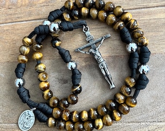 Puede incluir: Un rosario con cuentas de ojo de tigre marrón, nudos de cordón negro y cuentas de metal plateado pulido. La pieza incluye un crucifijo plateado con las letras INRI grabadas en la parte superior y un pequeño colgante ovalado de metal.