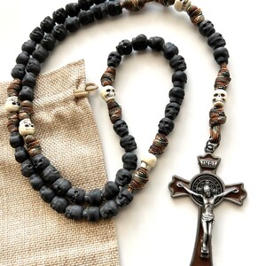 May include: A black and white skull rosary with a brown and silver Saint Benedict crucifix. The rosary is made with black beads and white skull beads. The crucifix has a brown background and a silver INRI inscription.