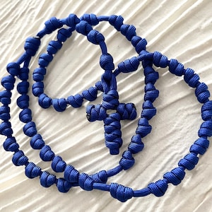 Puede incluir: Un rosario de cuerda azul anudado con una cruz anudada. El rosario está hecho de una cuerda gruesa y trenzada y tiene un diseño simple y elegante.