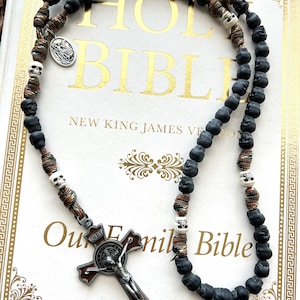 May include: A black and brown rosary with skull beads and a silver St. Francis of Assisi medal. The rosary is lying on a white book with the words "Holy Bible" and "New King James Version" printed on the cover.