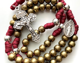 Może przedstawiać: Różaniec w kolorze czerwonym i złotym z srebrnym krucyfiksem i srebrnym medalikiem Matki Boskiej. Krucyfiks ma napis "JESUS NAZARENUS REX JUDAEORUM". Medalik ma napis "O Maryjo Niepokalanie Poczęta Módl się za nami".