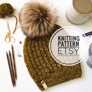 May include: A mustard yellow knitted beanie with a large brown faux fur pom pom. The beanie is made with a textured stitch and has a brown leather tag attached. The image also includes knitting needles, yarn, and a mug with the word "JOY" on it. The text "Knitting Pattern Etsy Aspenleafknits" is superimposed on the image.