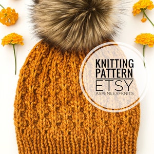 Puede incluir: Un gorro de punto amarillo mostaza con un gran pompón de piel sintética marrón. El gorro tiene un patrón de punto de cable. El texto "Knitting Pattern Etsy Aspenleafknits" está en un círculo blanco.