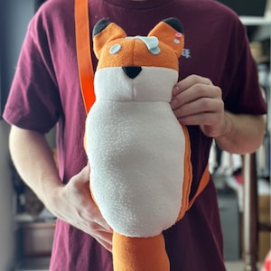 Könnte beinhalten: Ein Plüsch-Fuchs-Spielzeug mit orangefarbenem und weißem Fell, schwarzen Augen und einer schwarzen Nase. Der Fuchs trägt einen hellblauen Fleck auf dem Kopf und hat einen schwarzen Schwanz. Der Fuchs wird von einer Person in einem maroonfarbenen Hemd gehalten.