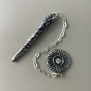 May include: A silver chain necklace with a textured silver pendant shaped like a long, thin, pointed object and a round, textured silver pendant with a clear rhinestone in the center.