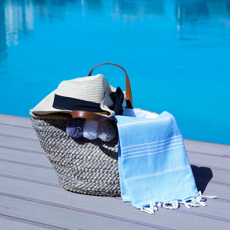 May include: A woven straw beach bag with a brown leather handle and a white and blue striped towel. The bag is filled with a straw hat and white pom-poms.
