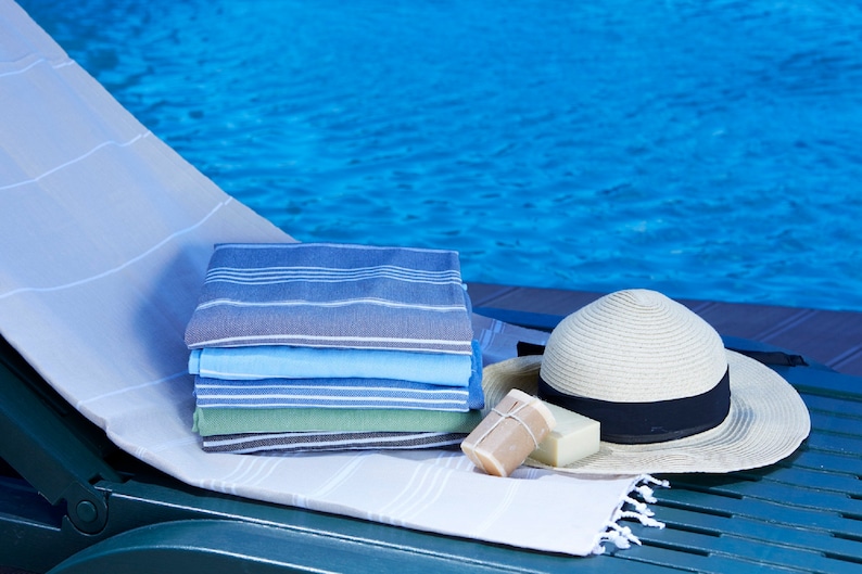 May include: A stack of striped towels in blue, green, and gray colors, a straw hat with a black band, and two bars of soap on a white towel on a green lounge chair by a pool.