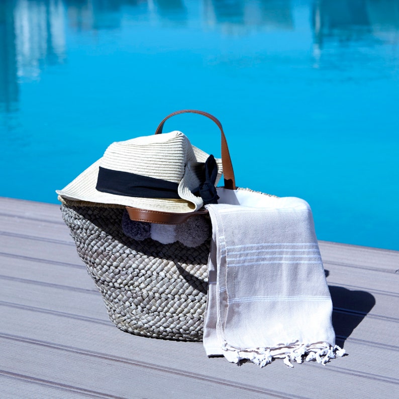 May include: A woven straw beach bag with a brown leather handle and a white straw hat with a black ribbon. A white and gray striped beach towel is hanging out of the bag.