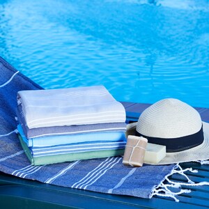 May include: A stack of folded towels in various shades of blue, green, and gray, with a straw hat and two bars of soap, on a green wooden lounge chair by a swimming pool.