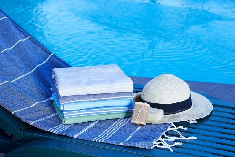 May include: A stack of folded towels in various shades of blue, green, and gray, with a straw hat and two bars of soap, on a green wooden lounge chair by a swimming pool.