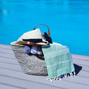 May include: A woven straw beach bag with a brown leather handle, a straw hat, a green and white striped towel, and white pom-poms.