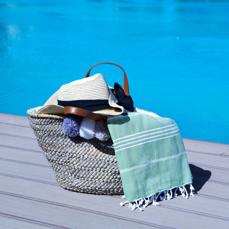 May include: A woven straw beach bag with a brown leather handle, a straw hat, a green and white striped towel, and white pom-poms.