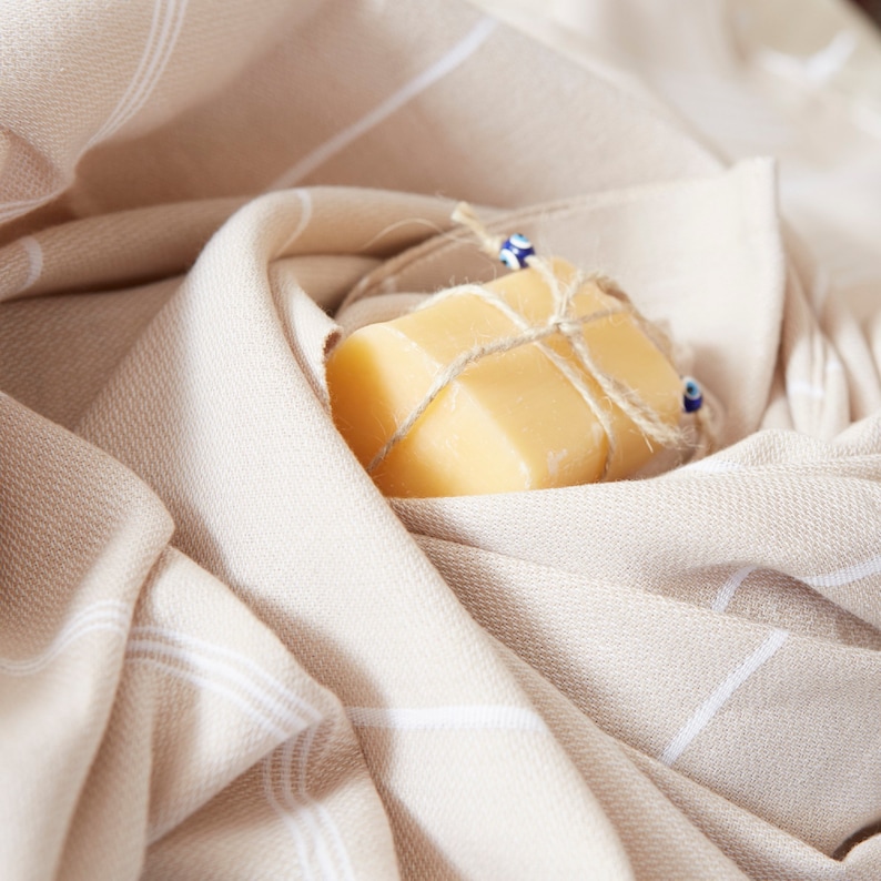 May include: A bar of soap wrapped in twine and decorated with blue and white beads, nestled in a beige and white striped fabric.