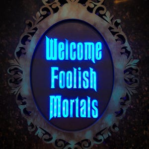 May include: An oval sign with the text "Welcome Foolish Mortals" in bright blue. The sign has a distressed, ornate frame with a teal-green finish, creating a vintage aesthetic. The dark background makes the glowing text stand out.