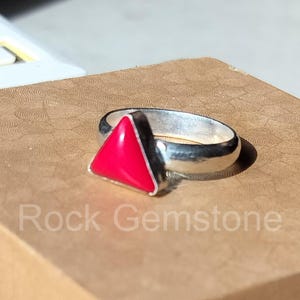 Puede incluir: Un anillo de plata con una gema triangular de color rojo brillante. El anillo está sobre una superficie marrón con el texto "Rock Gemstone". La gema es un punto focal vibrante.