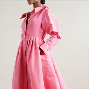 May include: A long pink dress with a collared neckline and long sleeves. The dress has a fitted bodice and a flowing skirt.
