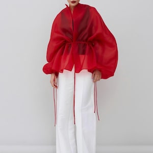 May include: A woman's fashion ensemble featuring a sheer red top with voluminous sleeves and a cinched waist, paired with wide-leg white pants. The outfit is completed with black shoes and red accents.