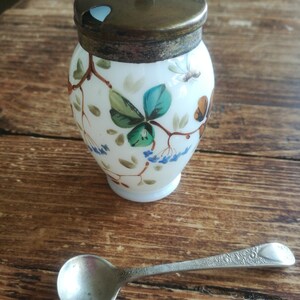 May include: A white ceramic sugar bowl with a floral design and a brass lid. The bowl is accompanied by a silver teaspoon.