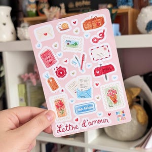 May include: A pink sticker sheet featuring illustrations of postal items, including stamps, envelopes, and a mailbox. The design includes hearts and a snail, with the words "Lettre d'amour".
