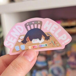 Peut inclure: Un autocollant rose avec les mots "SEND HELP!" présente une illustration de dessin animé d'une créature avec sa tête sur un bureau. Le bureau contient du matériel d'art, dont de la peinture, des crayons et des pinceaux. Deux petits gobelets bleus sont également sur le bureau.
