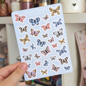 Peut inclure: Une planche d'autocollants avec divers motifs de papillons dans des tons de rose, de bleu, de jaune et de noir. Les autocollants sont sur un fond bleu clair, avec le texte "Whimsical Butterflies" en haut. Les papillons ont des motifs d'ailes détaillés.
