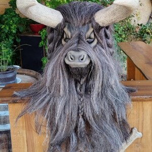 May include: A grey and brown furry mask with horns and a braided beard. The mask has a bull-like face with a black nose and closed eyes.