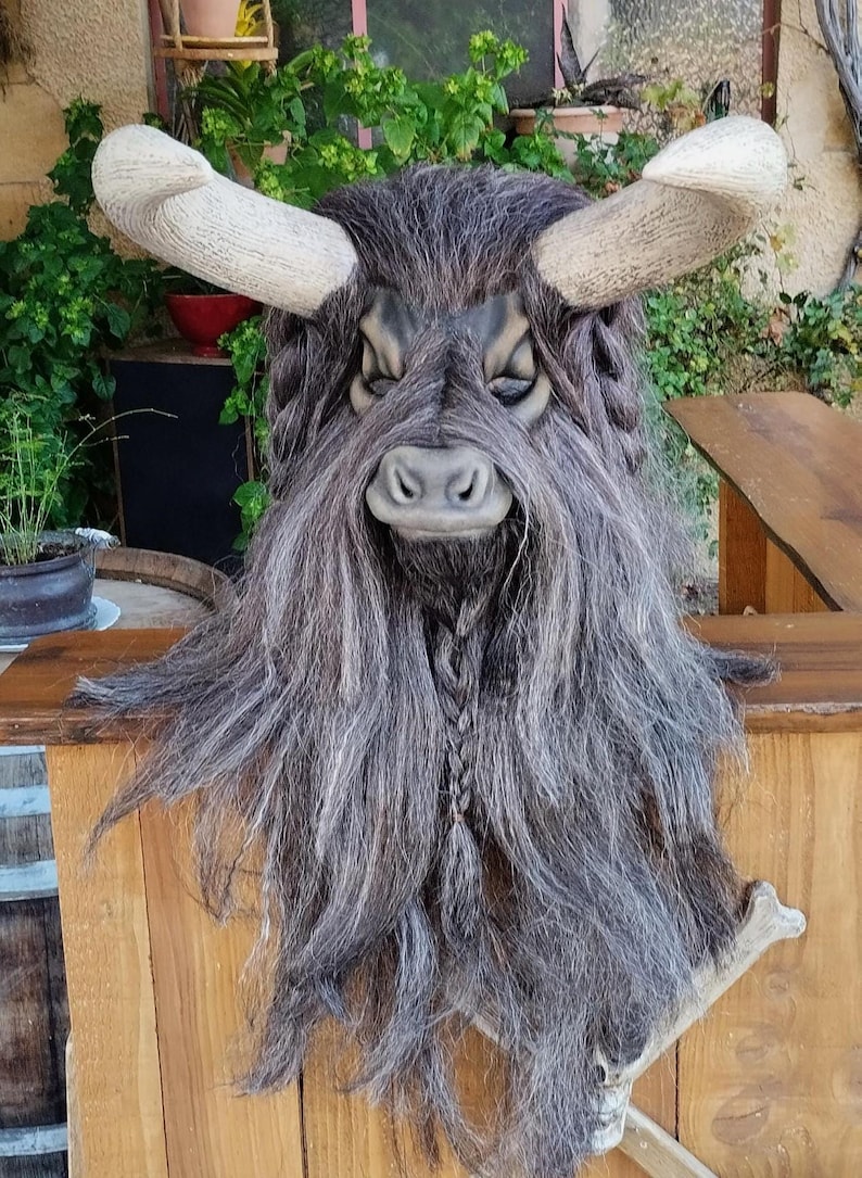 May include: A grey and brown furry mask with horns and a braided beard. The mask has a bull-like face with a black nose and closed eyes.