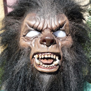 May include: A close-up of a werewolf mask with a snarling expression. The mask is made of brown latex and has realistic fur. The eyes are white and the teeth are sharp.