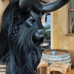 May include: A black bull mask with long, curly hair and horns. The mask is sitting on a wooden table with a barrel and stools in the background.