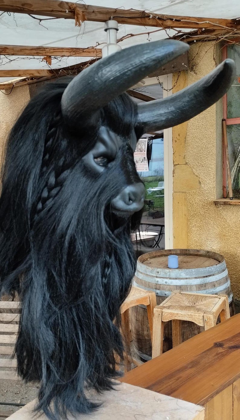 May include: A black bull mask with long, curly hair and horns. The mask is sitting on a wooden table with a barrel and stools in the background.