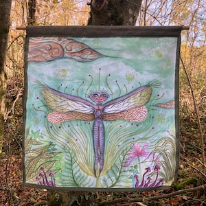 Op de afbeelding: Een groene wandkleed met een aquarel schilderij van een libel met een menselijk gezicht. De libel is omringd door groene en paarse planten en bloemen. De wandkleed hangt aan een boom in een bos.