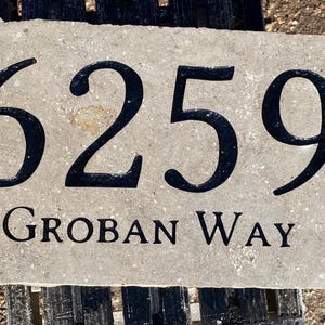 May include: A beige stone house number sign with black lettering. The number is 6259 and the street name is Groban Way.