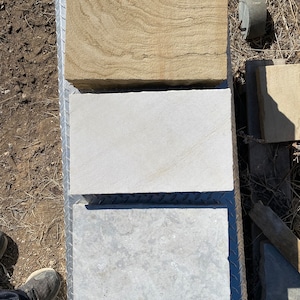 May include: Three rectangular stone tiles, one light brown, one white, and one light gray, are stacked on top of each other on a metal surface.