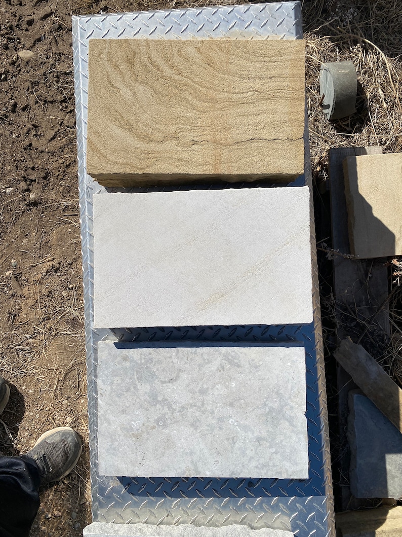 May include: Three rectangular stone tiles, one light brown, one white, and one light gray, are stacked on top of each other on a metal surface.