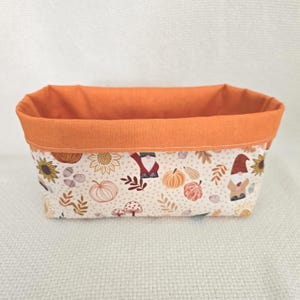 Op de afbeelding: Een rechthoekige stoffen opbergmand met een effen oranje bovenrand en een crèmekleurige basis met illustraties van kabouters, zonnebloemen, pompoenen, paddenstoelen en herfstbladeren. De mand heeft een gestructureerd ontwerp met open bovenkant.