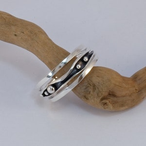 May include: A silver ring with a black band and silver beads. The ring is on a piece of driftwood.