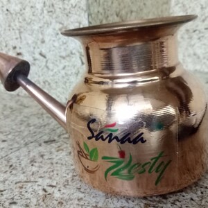 May include: A polished copper vessel with a wide, flared rim and a single narrow, angled spout featuring a conical tip. The body of the vessel displays the printed text Sanaa Zesty in dark blue and green, accompanied by a small mortar and pestle icon.