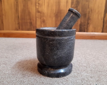 May include: A dark gray granite mortar and pestle set. The mortar is a cylindrical bowl with a rounded base. The pestle is a matching dark gray color and is resting inside the mortar. The set is designed for grinding herbs and spices.