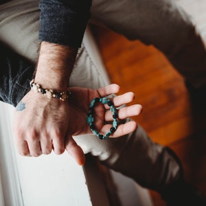 Könnte beinhalten: Zwei Armbänder mit Perlen. Ein Armband hat goldfarbene Perlen und schwarze Akzente. Das andere Armband hat türkisfarbene und schwarze Perlen. Die Armbänder werden auf den Händen einer Person präsentiert.