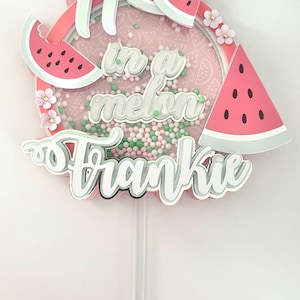 Könnte beinhalten: Ein rosa-grüner Tortendekor mit dem Text "1 in a melon Frankie" und Wassermelonen-Scheiben. Der Dekor ist auf weißem Hintergrund.