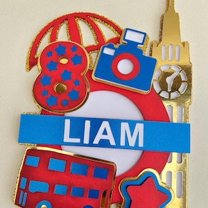 Op de afbeelding: Een rode, witte en blauwe taarttopper met een nummer 8, een camera, een dubbeldekkerbus, een ster, een paraplu en Big Ben. De topper heeft ook een blauwe banner met de naam "LIAM" erop.