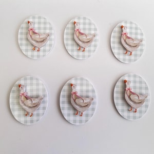 May include: Six oval-shaped stickers featuring a watercolor illustration of a goose wearing a pink bow. The geese are set against a light gray and white checkered background. The geese have orange beaks and feet.