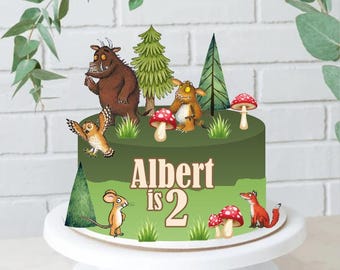 May include: A green birthday cake decorated with cartoon characters, trees, and mushrooms. The cake has the text "Albert is 2" in white. The cake is on a white cake stand.