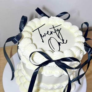 Op de afbeelding: Een witte taart met twee lagen, witte frosting en zwarte lintaccenten. De bovenkant van de taart heeft de woorden "Twenty One" in zwart geschreven. De taart staat op een wit bord.