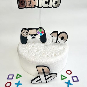 Puede incluir: Un conjunto de adornos para tartas que incluye un mando de PlayStation plateado, el logotipo de PlayStation y el número 10. El conjunto también incluye una pancarta plateada con el nombre "Benicio" y cuatro símbolos de botones de PlayStation de colores.